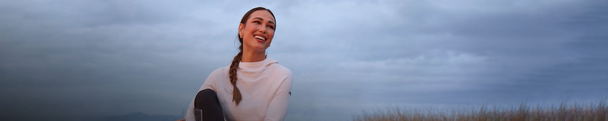 A woman wearing a KJUS Kessy Hooded Sweater smiles with a cloudy backdrop behind her.