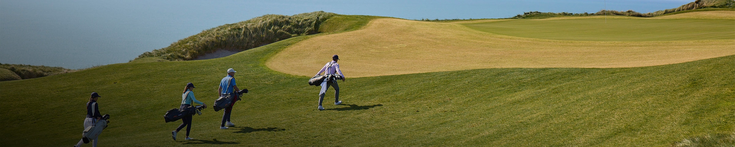 Four golfers carrying their clubs walk up to the green from the fairway.