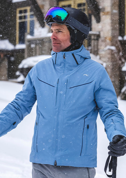 A male skier standing on a snowy ski mountain wearing a black helmet with goggles and a blue KJUS zip-up ski jacket.