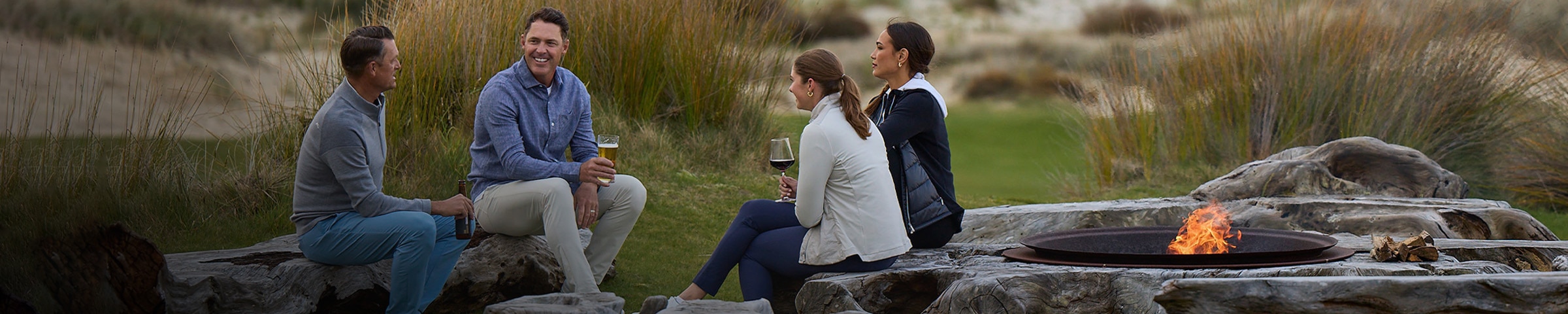 A group of people sit around a fire pit near a golf course, chatting and sipping beverages.