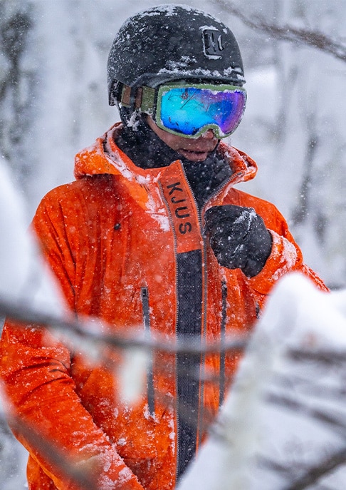 A male skier zipping up his orange KJUS ski jacket standing in snowy trees wearing a black helmet with goggles.