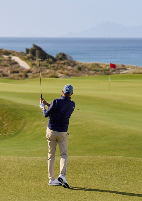 A male golfer finishing his golf swing wearing a white KJUS polo and black KJUS pants standing in front of a blue sky.