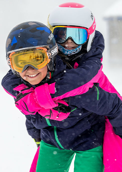 A junior skier wearing navy and green KJUS ski outfit carrying young female skier on his back, who is wearing a navy and pink KJUS ski outfit. They are standing on a snowy mountain background.