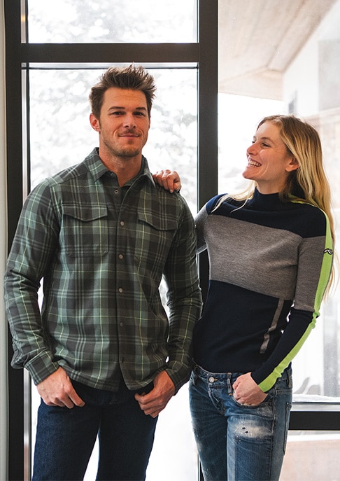 A man and a woman wearing KJUS lifestyle clothing link arms in front of a window displaying a snowy backdrop.