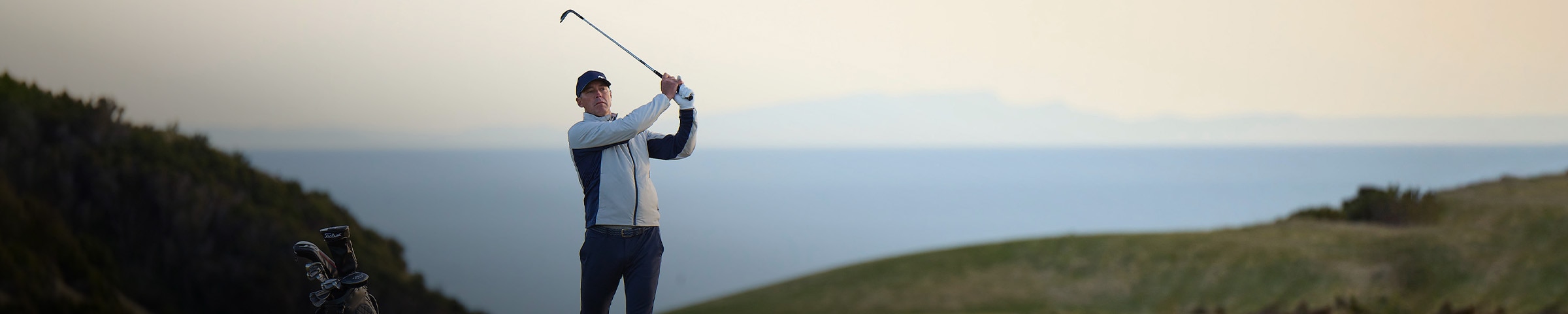 A male golfer in a KJUS Radiation Jacket swings a wedge on the fairway at sunrise.