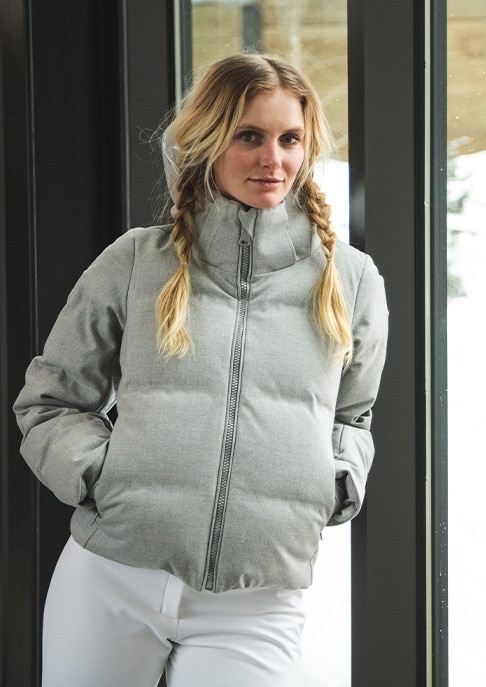 A woman wearing the Artic Luxe Jacket and KJUS ski pants leans on a window showing a snowy backdrop.