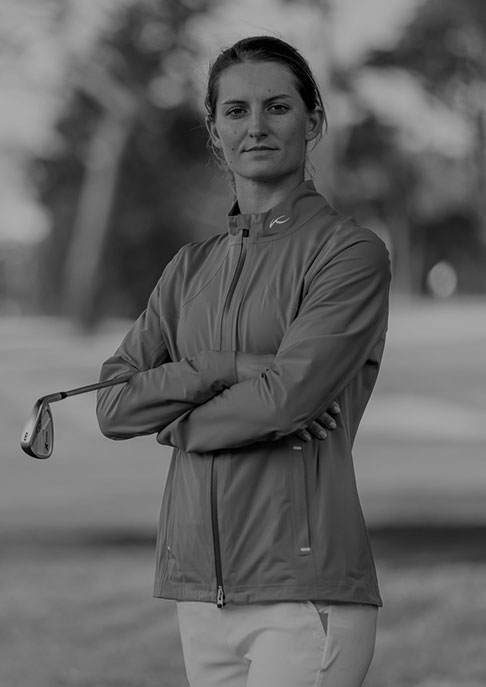 A female golfer wearing a KJUS rain jacket standing with her arms crossed and holding a golf club under one arm, posing on a golf course.