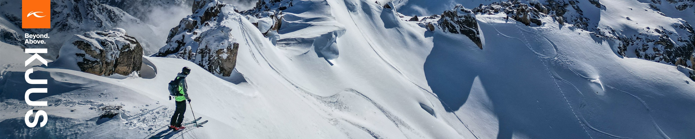 A skier wearing a KJUS jacket stands on a snowy mountaintop, looking down at the epic ski run before him.