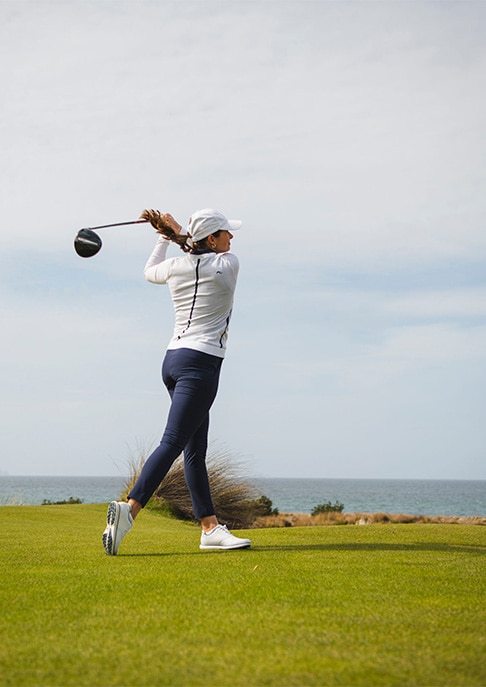 A women golfer finishing her swing on a green golf course, wearing a white KJUS jacket and KJUS pants.