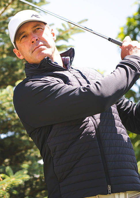 A male golfer finishing his golf swing wearing a white KJUS cap and a black KJUS zip-up insulation jacket standing in front of greenery.