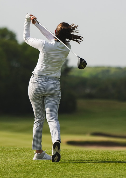 A women golfer finishing her swing on a green golf course, wearing a white KJUS polo and KJUS pants.