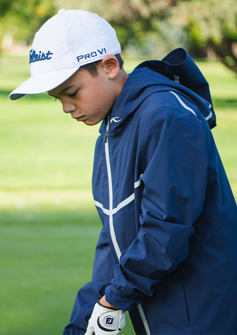 Two boys laugh on a golf course, one holding a club, the other a golf ball, both wearing golf caps and KJU golf jackets.