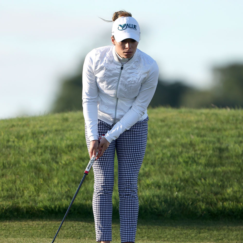 KJUS athlete Gaby Lopez, wearing a cap, white jacket, and checkered pants, is putting on a green golf course under a clear sky.