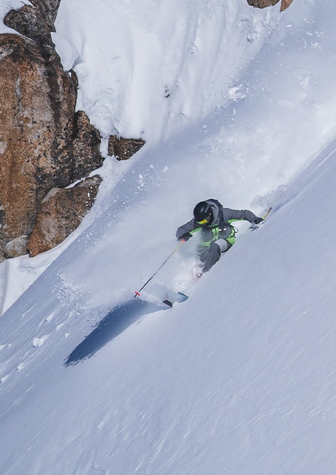 A skier wearing a KJUS jacket glides down a steep slope, kicking up snow around him.