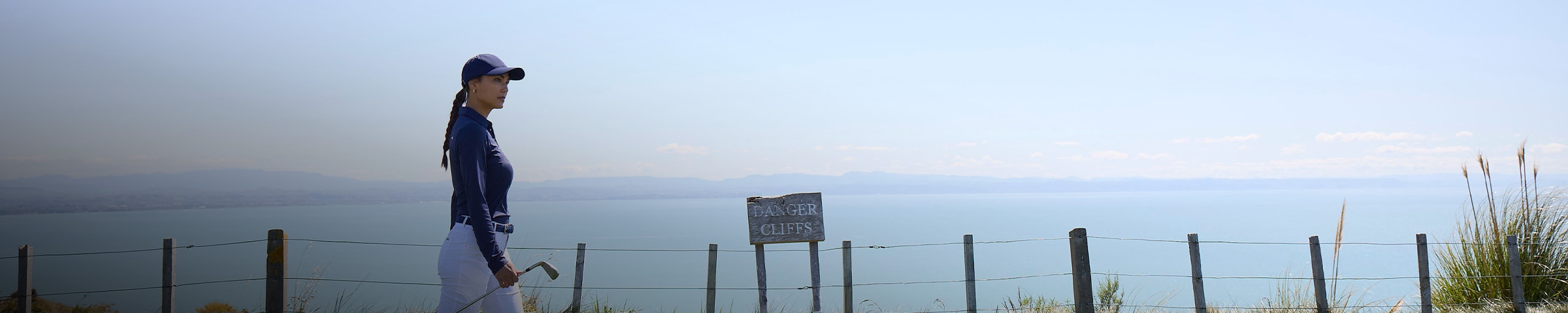 A woman wearing a KJUS golf polo carries a wedge to the next hole, a wire fence behind her reads 'DANGER CLIFFS.'