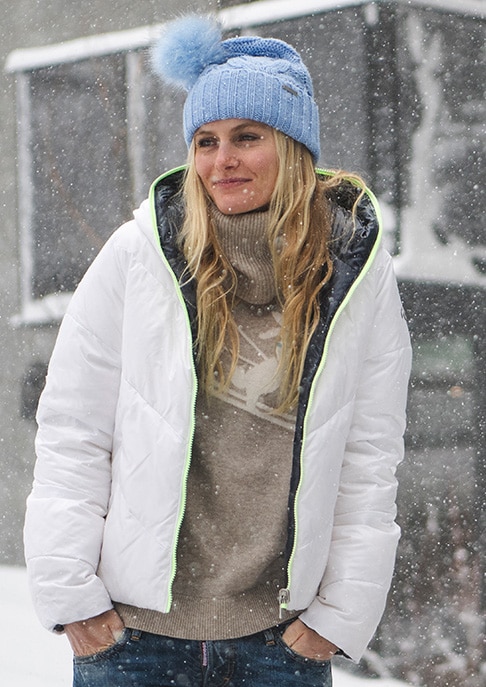 A smiling woman standing in the snowy outdoors wearing a KJUS puffer jacket, sweater, and beanie.