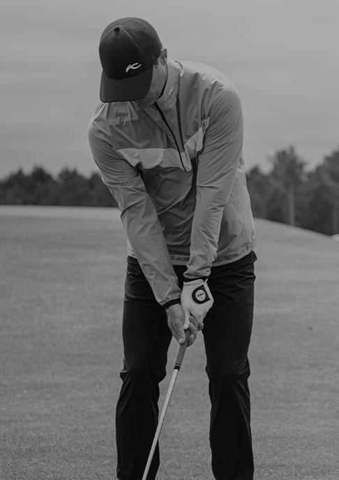 A golfer is preparing to swing his club wearing a KJUS rain jacket, long pants and standing on a golf course.