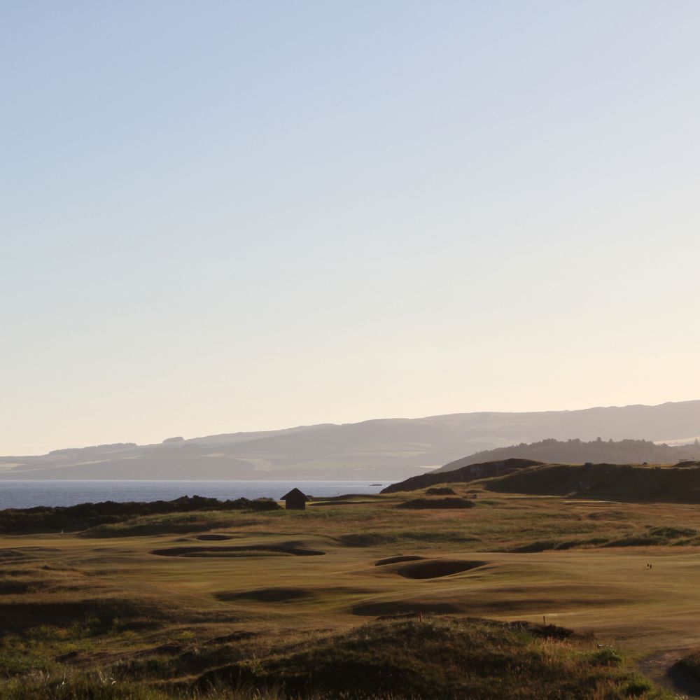 A grassy golf course with sand bunkers overlooking a calm sea, with rolling hills and a small hut in the background under a clear sky.