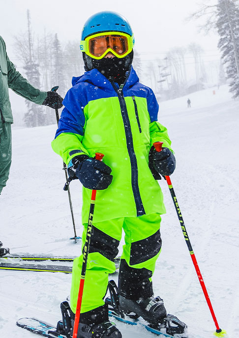 A junior skier wearing a bright green and blue KJUS ski outfit standing on a snowy mountain in his skis holding his ski poles.