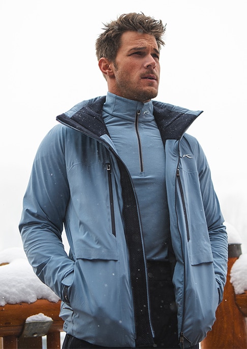 A man wearing the Lasse Airflow Jacket unzipped looks into the distance with a chairlift behind him.