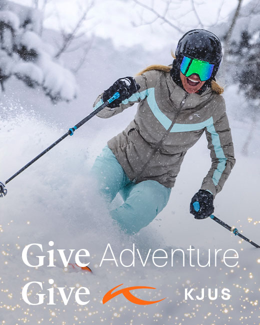 Female skier wearing tan and aqua blue ski jacket with aqua ski pants, skiing down a snowy run with text that reads 'Give Adventure Give KJUS' surrounded by holiday sparkles.