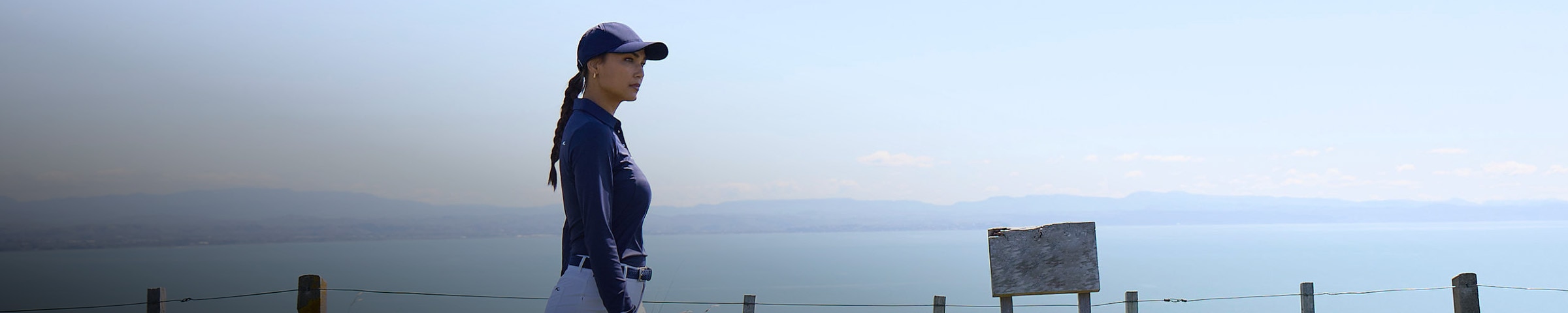 A woman wearing a KJUS polo shirt stands on a golf course with a large expanse of ocean behind her.