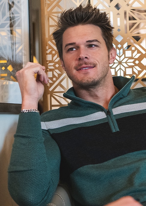 A man in a KJUS green stripe sweater sitting in a chair with a gold winter themed wall behind him.