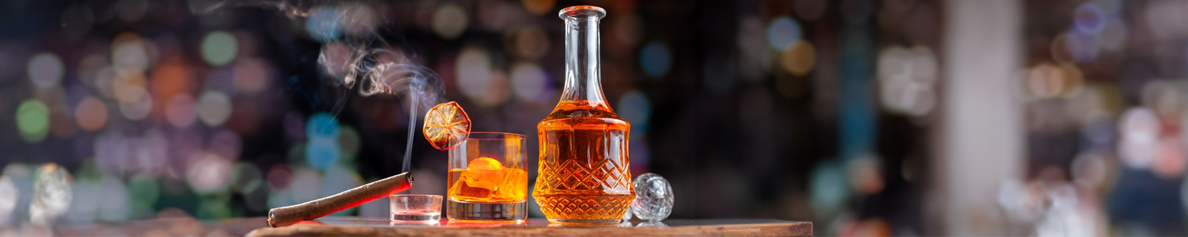 A glass of amber liquid (whiskey) with ice cubes sits on a dark wooden surface against a dimly-lit, blurred background.