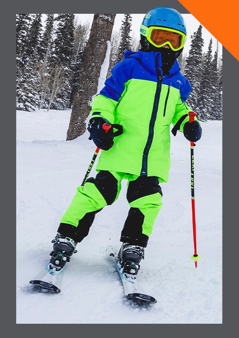 A junior skier wearing a bright green and blue KJUS ski outfit standing on a snowy mountain in his skis holding his ski poles.