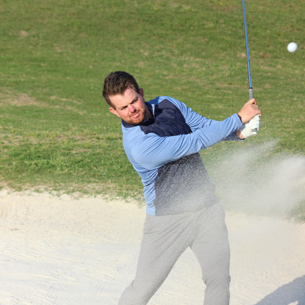 KJUS athlete Morgan Deneen swings a golf club, hitting a ball out of a sand bunker, with green grass surrounding the sandy area.