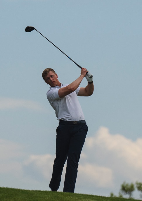 A male golfer finishing his golf swing wearing a white KJUS polo and black KJUS pants standing in front of a blue sky.