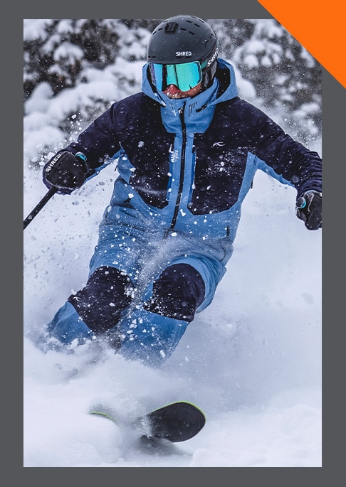 A male skier zipping up his orange KJUS ski jacket standing in snowy trees wearing a black helmet with goggles.