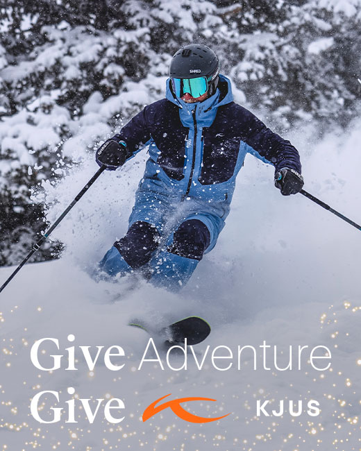 Skier in snow wearing navy and blue zip-up ski jacket and matching pants with shiny goggles and a black helmet surrounded by snowy trees with text that reads 'Give Adventure Give KJUS' and holiday sparkle details.