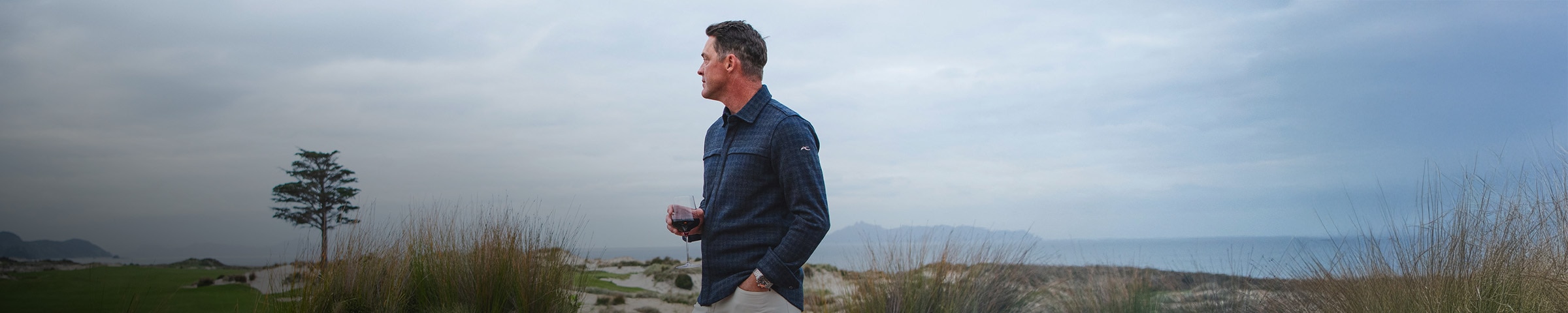 A man wearing a KJUS Yellowstone Shirt holds a glass of wine and looks out over the course from the clubhouse.