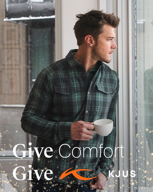 Man wearing plaid button-up shirt with pockets in a green and navy color, holding his coffee cup and glancing out the window with text that reads 'Give Comfort Give KJUS' and has holiday sparkles around the text.