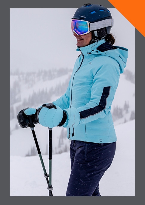 A smiling woman standing in the snowy outdoors wearing a KJUS puffer jacket, sweater, and beanie.